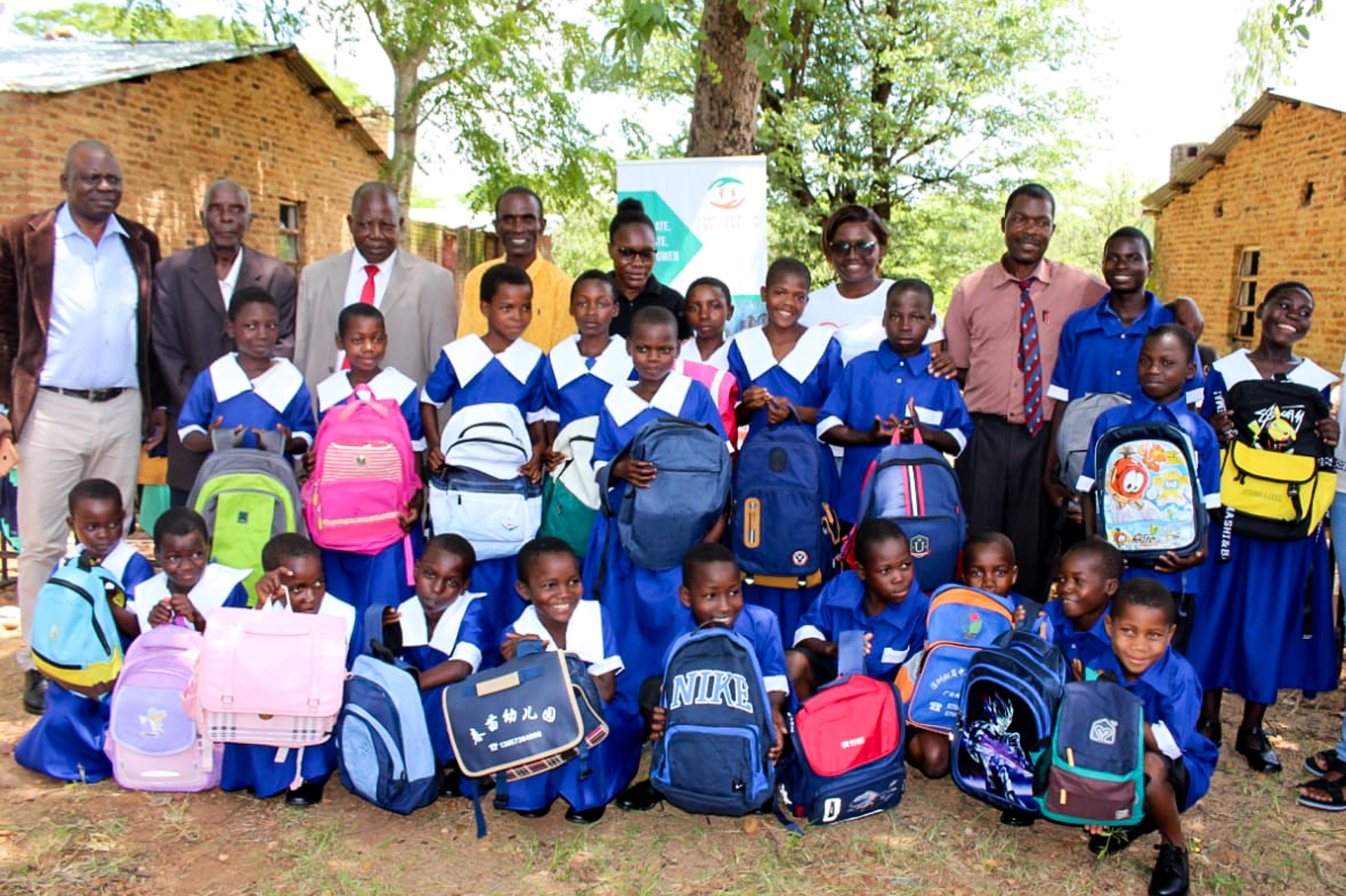 Ein großes Gruppenfoto im Freien zeigt eine Gruppe von Schulkindern aus Malawi in blau-weißen Uniformen, die stolz ihre neuen, bunten Rucksäcke präsentieren. Die Kinder knien in der vorderen Reihe oder stehen dahinter, umgeben von mehreren Erwachsenen, die ebenfalls lächeln. Im Hintergrund sind einfache Backsteingebäude und Bäume unter einem hellen Himmel zu sehen.