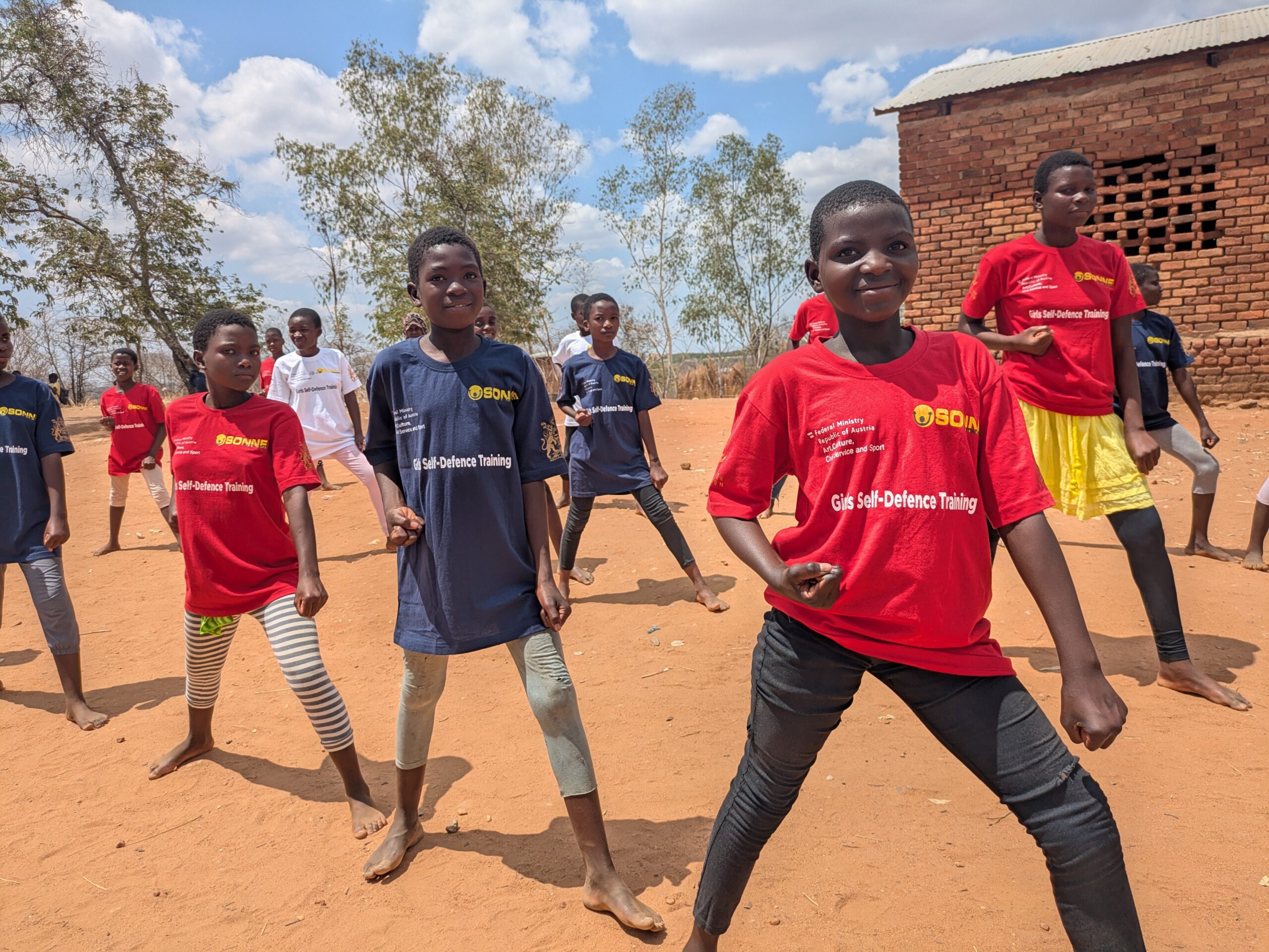 Malawi, Selbstverteidigung Eine Gruppe junger Mädchen übt Selbstverteidigung auf einem sandigen Platz im Freien. Sie stehen in einer entschlossenen Kampfkunst-Pose mit gebeugten Knien und geballten Fäusten. Die Mädchen tragen rote und dunkelblaue T-Shirts mit dem Logo von SONNE-International und der Aufschrift „Girls Self-Defence Training“. Im Hintergrund sind ein Backsteingebäude und Bäume unter einem leicht bewölkten Himmel zu sehen.