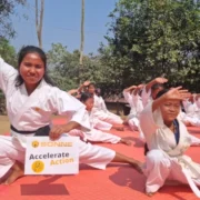 Eine junge Frau im weißen Karate-Anzug mit schwarzem Gürtel hockt lächelnd im Vordergrund auf einer roten Matte. Sie hält ein weißes Schild mit dem Logo von SONNE-International und der Aufschrift „Accelerate Action“ in die Kamera. Neben ihr hockt ein jüngeres Mädchen in ähnlicher Pose; im Hintergrund trainiert eine größere Gruppe von Kindern in Karate-Uniformen unter freiem Himmel.