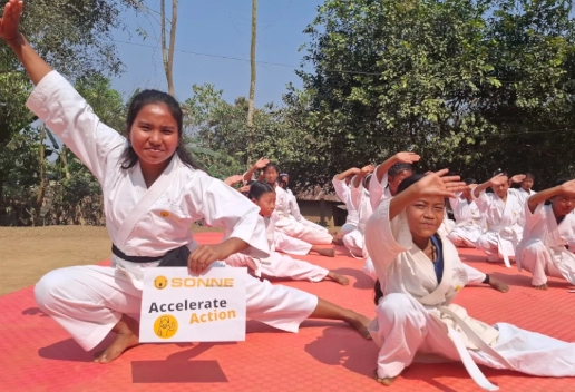 Eine junge Frau im weißen Karate-Anzug mit schwarzem Gürtel hockt lächelnd im Vordergrund auf einer roten Matte. Sie hält ein weißes Schild mit dem Logo von SONNE-International und der Aufschrift „Accelerate Action“ in die Kamera. Neben ihr hockt ein jüngeres Mädchen in ähnlicher Pose; im Hintergrund trainiert eine größere Gruppe von Kindern in Karate-Uniformen unter freiem Himmel.