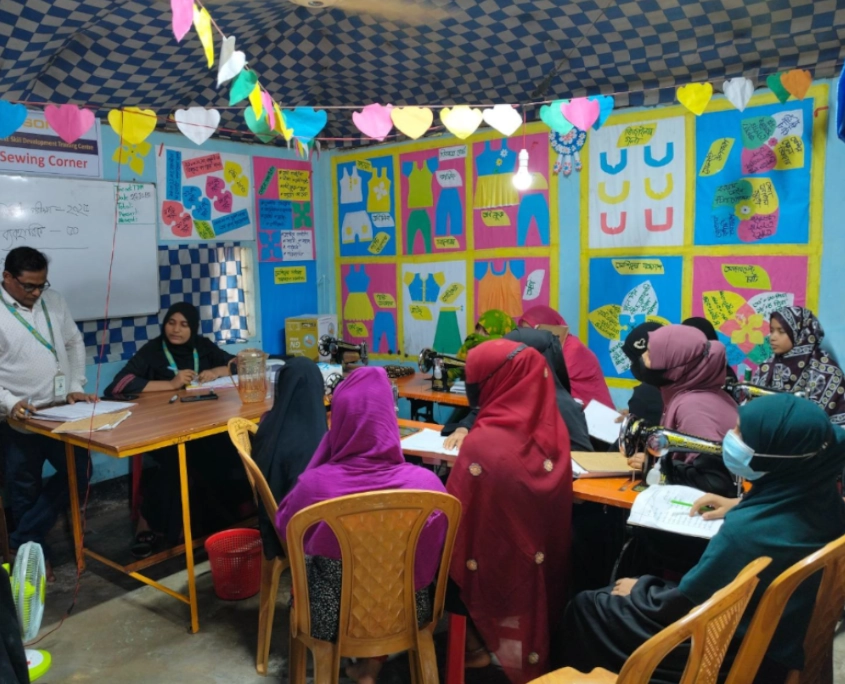 Bangladesch Rohingya Beitragsbild 3 Ein Schulungsraum mit blauen Wänden, die mit vielen bunten Papier-Schnittmustern von Kleidungsstücken beklebt sind. Im Raum sitzt eine Gruppe von Frauen mit bunten Kopftüchern an Tischen, vor sich Nähmaschinen und offene Hefte. Links steht ein männlicher Ausbilder mit Brille und Ausweis am Hemd, der zu der Gruppe spricht; neben ihm sitzt eine Frau an einem Schreibtisch. Von der Decke hängen bunte Papierherzen als Dekoration.