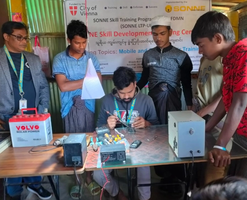 Bangladesch Rohingya Beitragsbild 5 Ein junger Mann sitzt an einem Tisch und lötet konzentriert an einem zerlegten Mobiltelefon. Um ihn herum stehen drei weitere junge Männer sowie ein älterer Mann mit Brille und einem Ausweis um den Hals, die ihm aufmerksam zusehen. Auf dem Tisch liegen diverse Werkzeuge, ein Messgerät, ein Netzteil und eine große Batterie mit der Aufschrift „Solar Power“. Im Hintergrund hängt ein weißes Banner mit den Logos der „City of Vienna“ und „SONNE-International“, das auf ein „Skill Training Program“ für „Mobile and Solar“ hinweist.