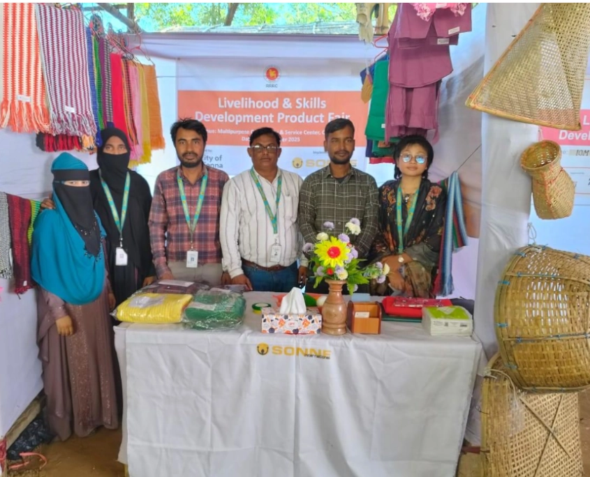 Bangladesch Rohingya Beitragsbild 6 Eine Gruppe von sechs Erwachsenen steht hinter einem Ausstellungsstand. Auf dem Tisch im Vordergrund liegen verpackte bunte Stoffe, Nähutensilien in einem Korb sowie eine Vase mit Kunstblumen; auf der Vorderseite des weißen Tischtuchs ist das Logo von SONNE-International zu sehen. Im Hintergrund hängt ein oranges Banner mit der Aufschrift „Livelihood & Skills Development Product Fair“. An den Seiten werden handgefertigte Produkte präsentiert: links hängen bunte Schals, rechts verschiedene geflochtene Bambuskörbe und Reusen.