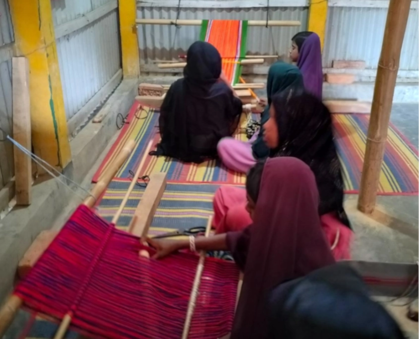 Bangladesch Rohingya Beitragsbild Eine Gruppe junger Frauen sitzt hintereinander auf bunten gestreiften Matten am Boden in einem Raum mit Wänden aus Wellblech und Bambus. Sie arbeiten konzentriert an einfachen, am Boden befestigten Webrahmen aus Holz. Vor ihnen sind leuchtend bunte Garne in Rot, Violett und Orange gespannt, aus denen sie Stoffe weben. Die Frauen tragen dunkle sowie farbige Kopftücher.
