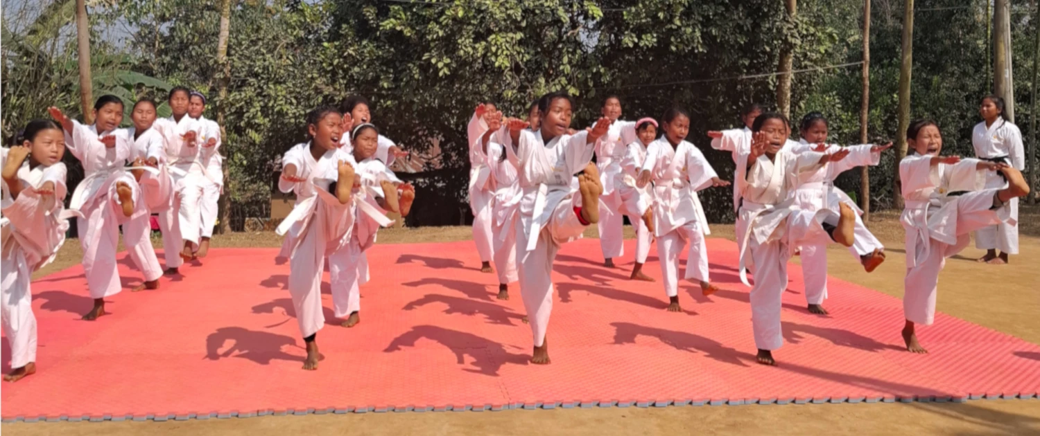 Bangladesch Sport Titelbild Eine große Gruppe junger Mädchen in weißen Karate-Uniformen trainiert im Freien auf einer großen roten Matte. Die Mädchen führen synchron einen hohen Tritt mit dem rechten Bein aus und haben dabei die Münder geöffnet, als würden sie energisch rufen. Im Hintergrund sind Bäume und eine ländliche Umgebung zu sehen.
