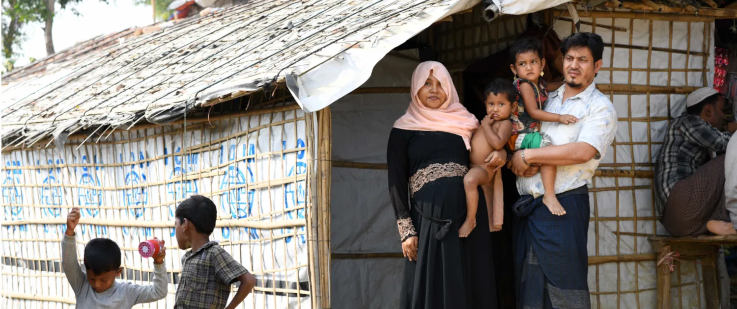 Bangladesch Top Slider Rohingya Eine Familie steht vor einer provisorischen Unterkunft, die aus Bambusstäben und weißen Plastikplanen errichtet wurde. Die Mutter trägt einen schwarzen Umhang und ein hellrosa Kopftuch und hält ein Kleinkind im Arm; neben ihr steht der Vater, der ebenfalls ein Kind trägt. Im linken Vordergrund sind zwei Jungen zu sehen, von denen einer ein Spielzeug hochhält.