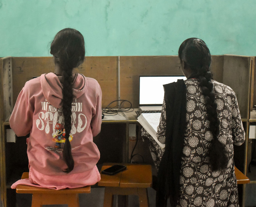 Indien-Computerkurs-1 Zwei Frauen sitzen in einem Computerkurs vor Bildschirmen. Man sieht sie von hinten.