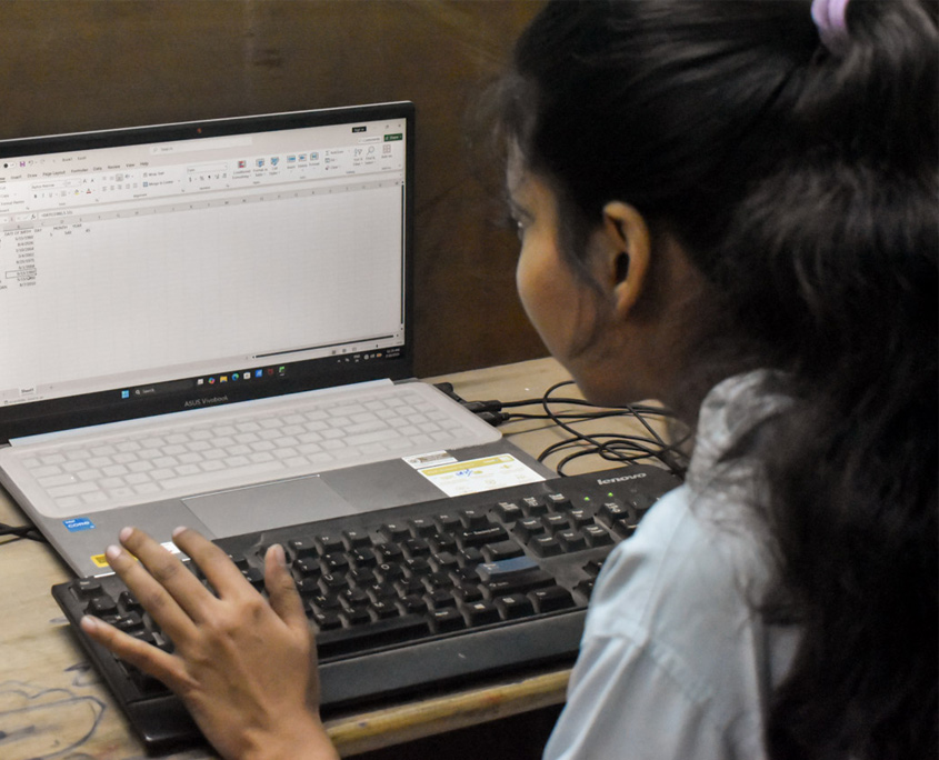 Indien-Computerkurs-3 Eine junge Frau sitzt in einem Computerkurs vor Bildschirmen. Man sieht sie von hinten.