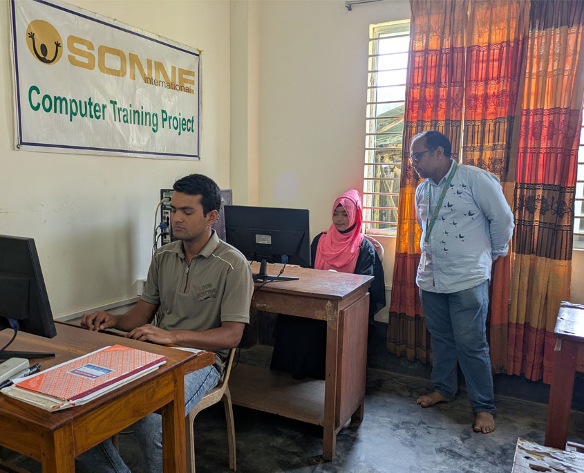 zwei studierende sitzen vor Computern in einer unterrichtsstunde. der lehrer unterstützt sie dabei. auf einem schild an der wand steht: SONNE computer training projec.t