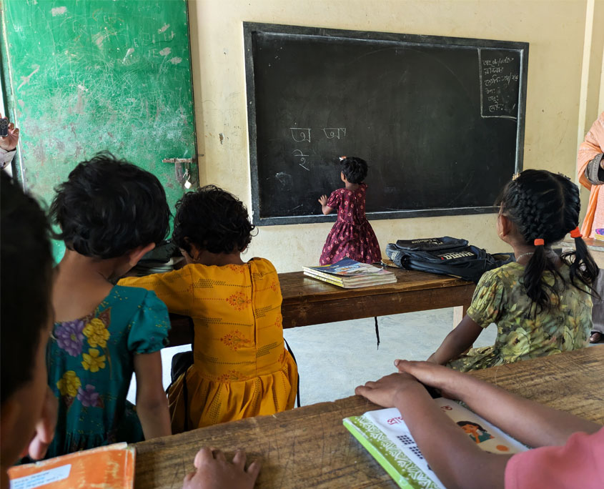 Ein Kind steht an der Tafel und schreibt auf Bangla. man sieht es von hinten.