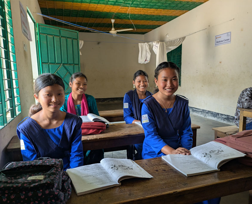 in einer Schule in Bangladesch sitzen vier jugendliche und lachen in die Kamera. sie sitzen auf Schulbänken und haben ihre Schulbücher geöffnet.