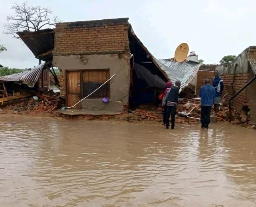 Familie vor ihrem zerstörten Haus in Malawi, Flutwasser und beschädigtes Dach im Hintergrund