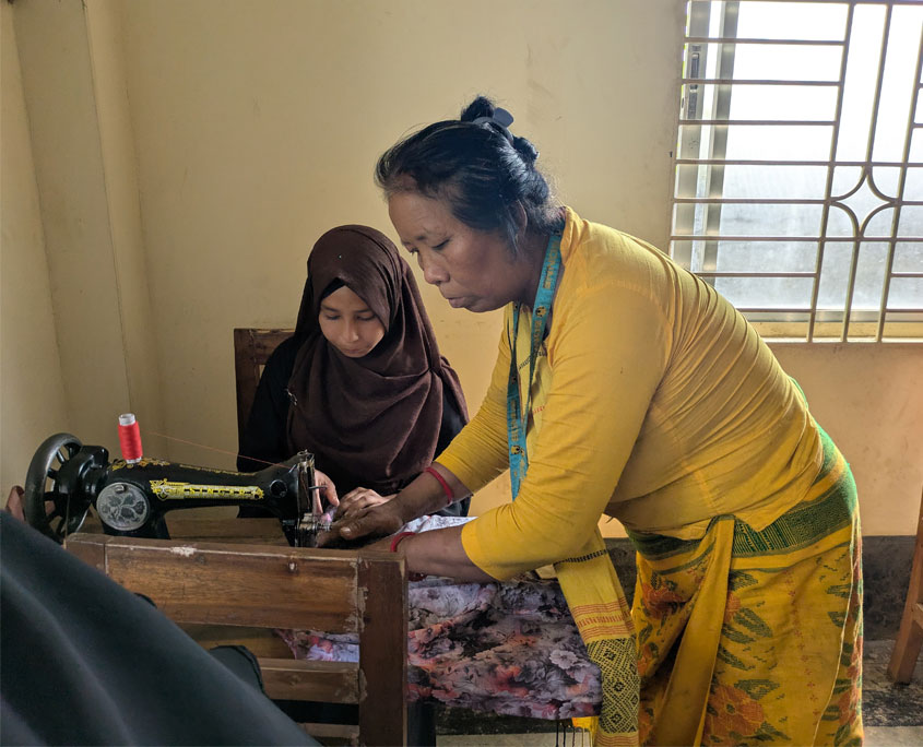 eine jugendliche sitzt vor einer Nähmaschine. die lehrerin zeigt ihr etwas dabei. Beim projektbesuch in Bangladesch