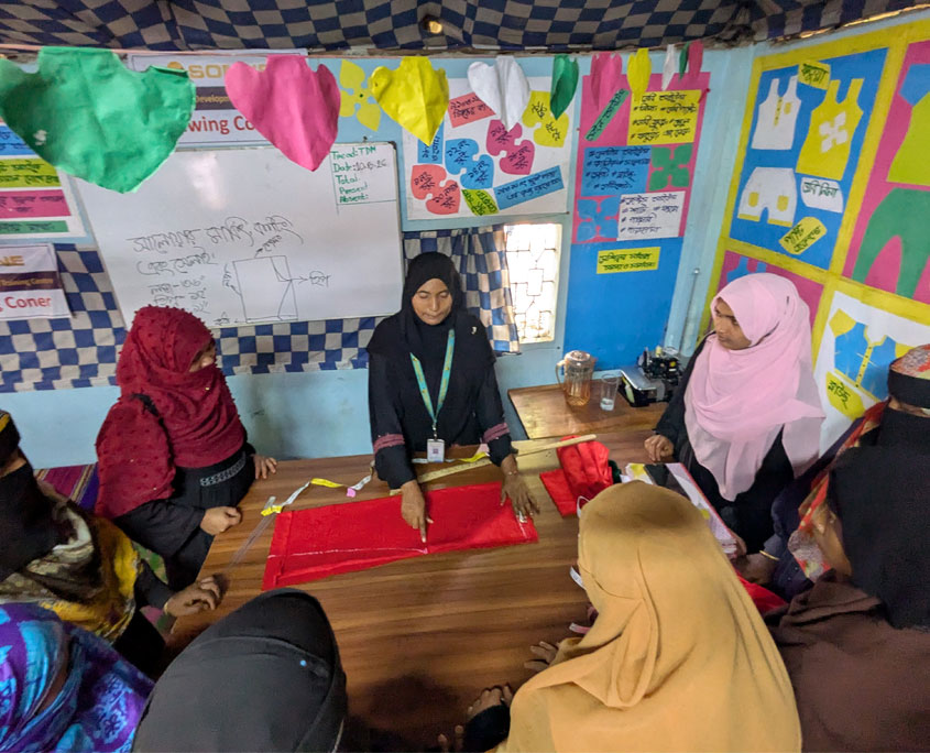 Das Bild zeigt ein Klassenzimmer in dem mehrere Frauen um einen Tisch stehen auf dem eine Trainierin gerade anleitet, wie man ein Schnittmuster auf den Stoff überträgt. Die Wände sind mit Plakaten von Schnittmustern bestückt. Bangladesch