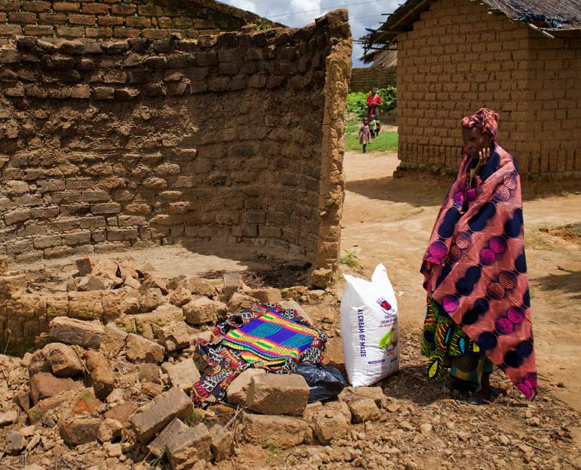 Malawi-Nothilfe-2 Eine Frau steht vor den Ruinen eines von der Flut zerstörten Hauses. Vor ihr ist ein großer Sack Maismehl, Decken und andere Hilfsgüter.