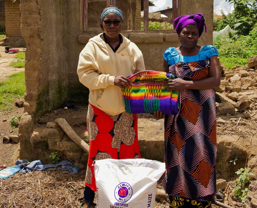 Malawi-Nothilfe-4 Eine Frau steht vor den Ruinen eines von der Flut zerstörten Hauses. Eine SONNE Mitarbeiterin überreicht ihr Lebensmittel und Decken