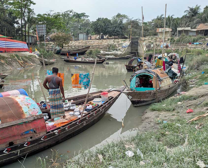 Mehrere traditionelle Holzboote liegen an einem schlammigen Flussufer auf der Insel Bhola. Auf einem der Boote steht ein Mann zwischen Metallgefäßen, während bunte Kleidung zwischen den Booten zum Trocknen hängt. Im Hintergrund stehen einfache Häuser und Bäume. Das Bild zeigt einen Moment aus dem Alltagsleben der Manta‑Gemeinschaft, die oft auf und entlang der Boote lebt.