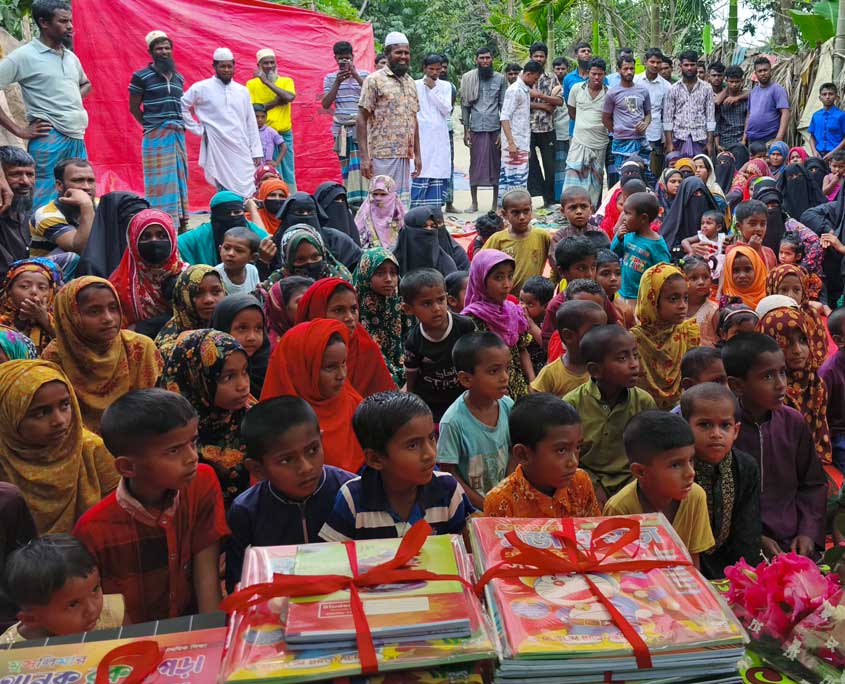 Schulkinder in Bangladesch sitzen am Boden, dahinter sitzen die Mütter und im Hintergrund stehen die Väter des Dorfes