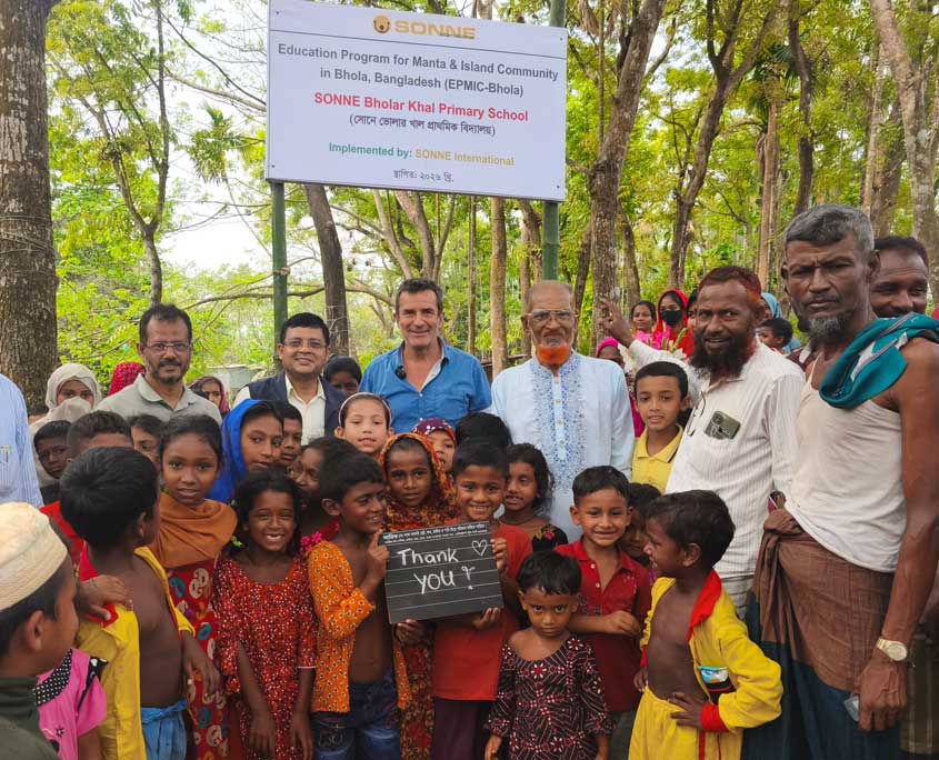 Im Vordergrund stehen Kinder der Manta Community und halten ein Schild mit "Thank You" in die Kamera. Dahinter stehen Mitarbetier von SONNE-International sowie ein Dorfvorsteher. Sie alle stehen vor einem Schild, das auf die baldige Öffnung einer SONNE-Schule hinweist.