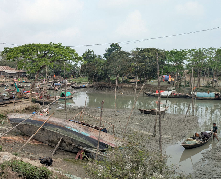 Mehrere kleine Holzboote liegen bei Ebbe auf einer schlammigen Uferzone eines Flussarms. Ein größeres Boot im Vordergrund ist zur Seite gekippt und von Holzpfählen umgeben. Menschen bewegen sich zwischen den Booten, einige arbeiten daran. Im Hintergrund stehen einfache Häuser, Bäume und aufgehängte Wäsche.