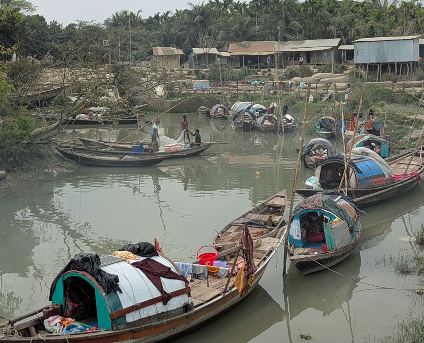 Holzboote der Manta‑Gemeinschaft liegen dicht an einem schmalen, trüben Fluss auf der Insel Bhola in Bangladesch. Viele Boote haben gebogene Planen- oder Blechdächer, unter denen Menschen sitzen oder arbeiten; Kleidung und Haushaltsgegenstände hängen oder stehen auf den Decks. Am Ufer stehen einfache Stelzenhäuser zwischen Bäumen.