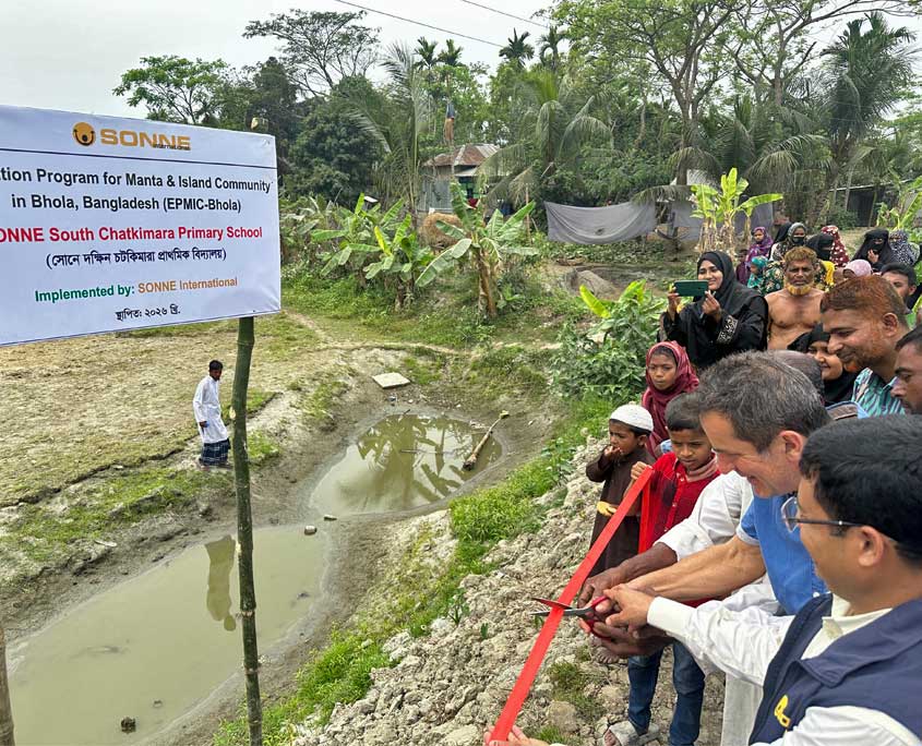 Die Einweihung des Schulgeländes, wo bald eine neue SONNE Schule entsteht wird gefeiert. Menschen stehen vor Grundstück zwischen Bananenstauden und Grün, während ein rotes Band zur Eröffnung einer neuen Schule für Kinder der Manta‑Gemeinschaft und anderer armer Familien auf der Insel Bhola in Bangladesch durchtrennt wird. Links steht ein Schild mit dem Namen des Bildungsprogramms und der Schule.