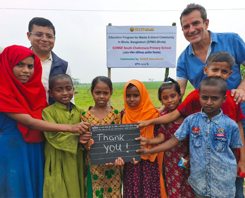 Eine Gruppe von Kindern und zwei Erwachsene stehen im Freien vor einem Schild der neuen Schule auf der Insel Bhola in Bangladesch. Die Kinder halten eine kleine Tafel mit der Aufschrift ‚Thank you!‘, im Hintergrund ist das Schild des Bildungsprogramms für die Manta‑ und Inselgemeinschaft zu sehen.