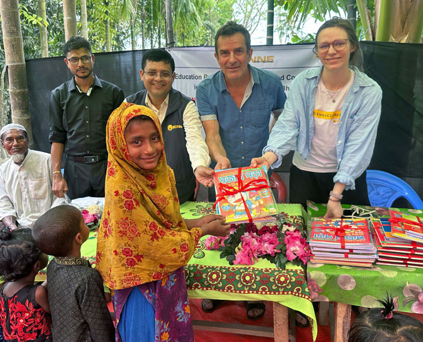 Ein Mädchen in bunter Kleidung und gelbem Kopftuch erhält ein Paket farbiger Bücher mit roter Schleife von mehreren Erwachsenen, die hinter einem Tisch mit weiteren Büchern und Blumen stehen. Die Gruppe befindet sich unter einem einfachen Sonnenschutz im Grünen. Das Bild zeigt eine Bücherausgabe im Rahmen des Bildungsprogramms für Kinder der Manta‑Gemeinschaft auf der Insel Bhola in Bangladesch.