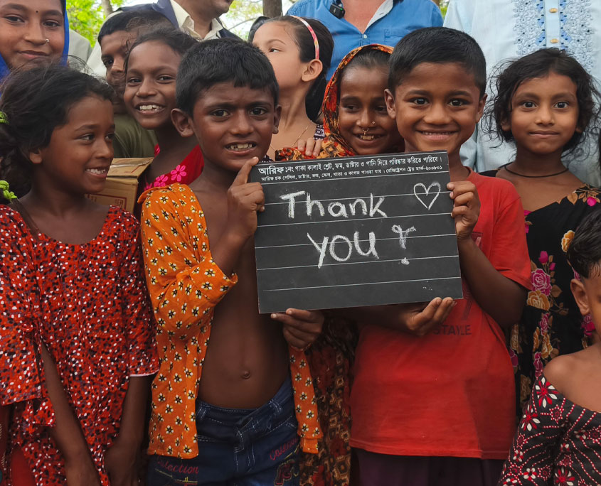 Manta-Kids-Thank-You Eine Gruppe von Kindern steht eng beisammen im Freien und lächelt in die Kamera. Zwei Kinder halten eine kleine Tafel mit der Aufschrift ‚Thank YOU!‘ und einem Herzsymbol. Hinter ihnen sind weitere Kinder und einige Erwachsene zu sehen. Das Foto zeigt einen Moment des Dankes im Rahmen des Schulprojekts für die Manta‑Gemeinschaft auf der Insel Bhola in Bangladesch