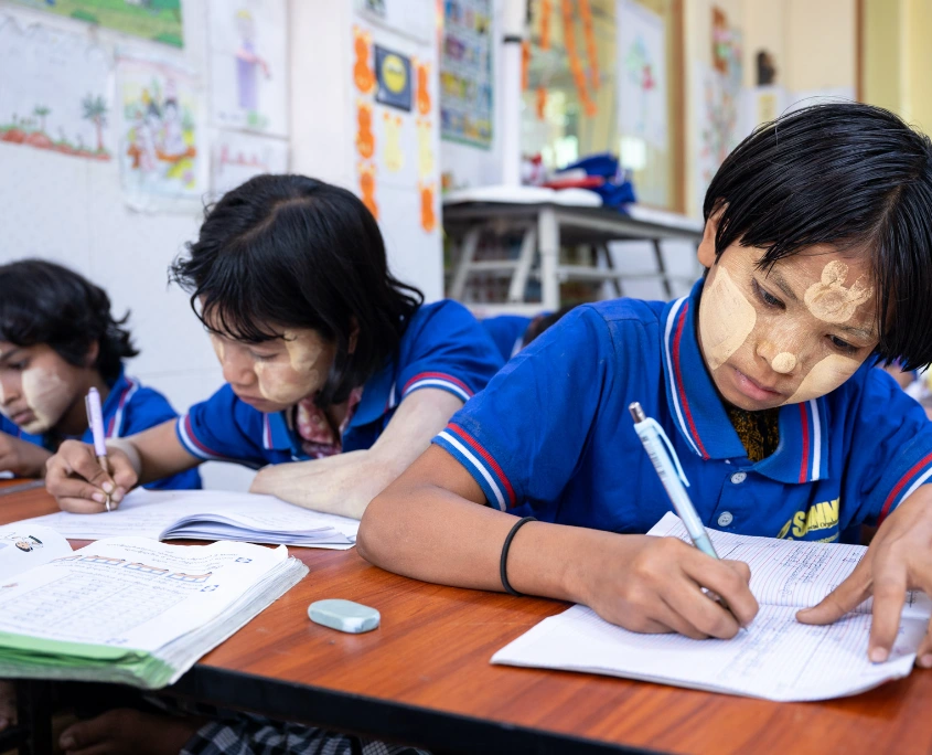 Drei Kinder mit SONNE-Shirts sitzen an einem Tisch und schreiben in ihre Hefte im Unterricht einer Tagesbetreuungsstätte in Yangon.
