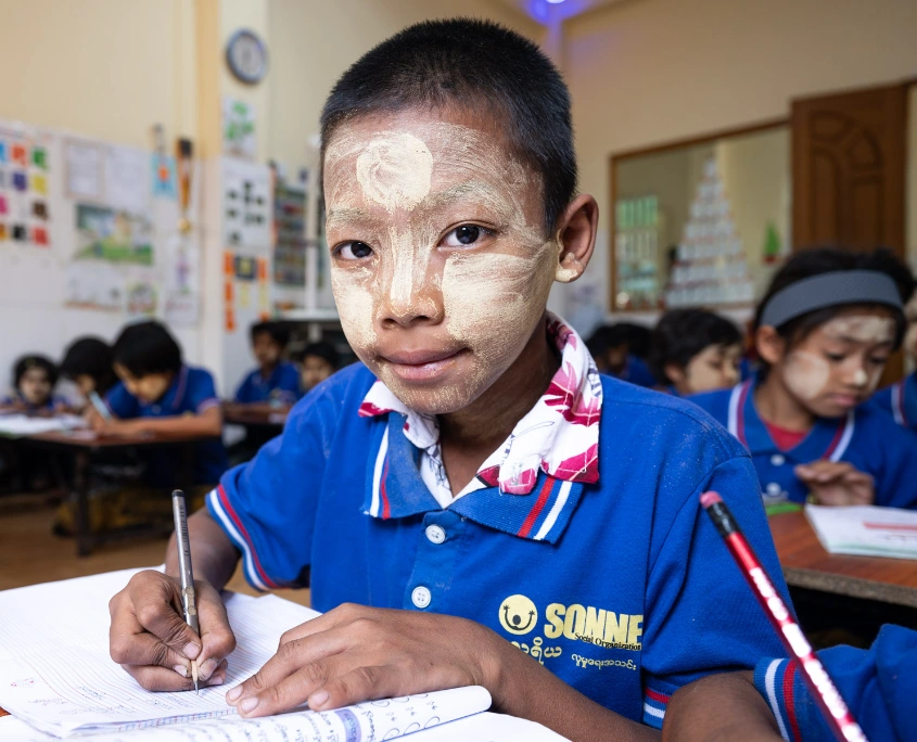 Bub mit Thanaka im Gesicht schreibt konzentriert in ein Heft in einer SONNE-Tagesbetreuungsstätte in Yangon, Myanmar.