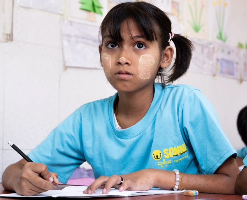 Mädchen in hellblauem SONNE-Shirt sitzt mit Stift und Heft im Klassenzimmer und lernt in einer SONNE-Tagesbetreuungsstätte in Myanmar.