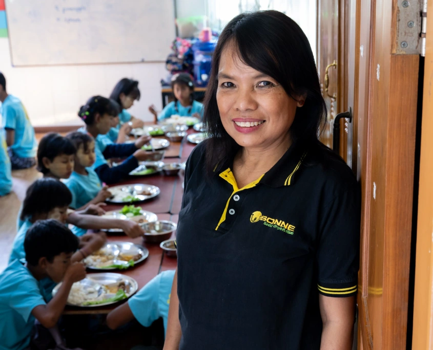 San San, Leiterin von SONNE Myanmar, steht lächelnd in einer Tagesbetreuungsstätte, während Kinder im Hintergrund gemeinsam essen.