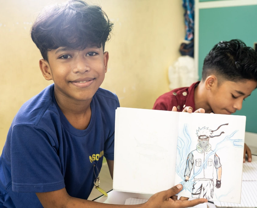Lächelnder Bub in blauem SONNE-Shirt zeigt stolz eine Zeichnung in seinem Heft, während ein weiteres Kind neben ihm arbeitet.