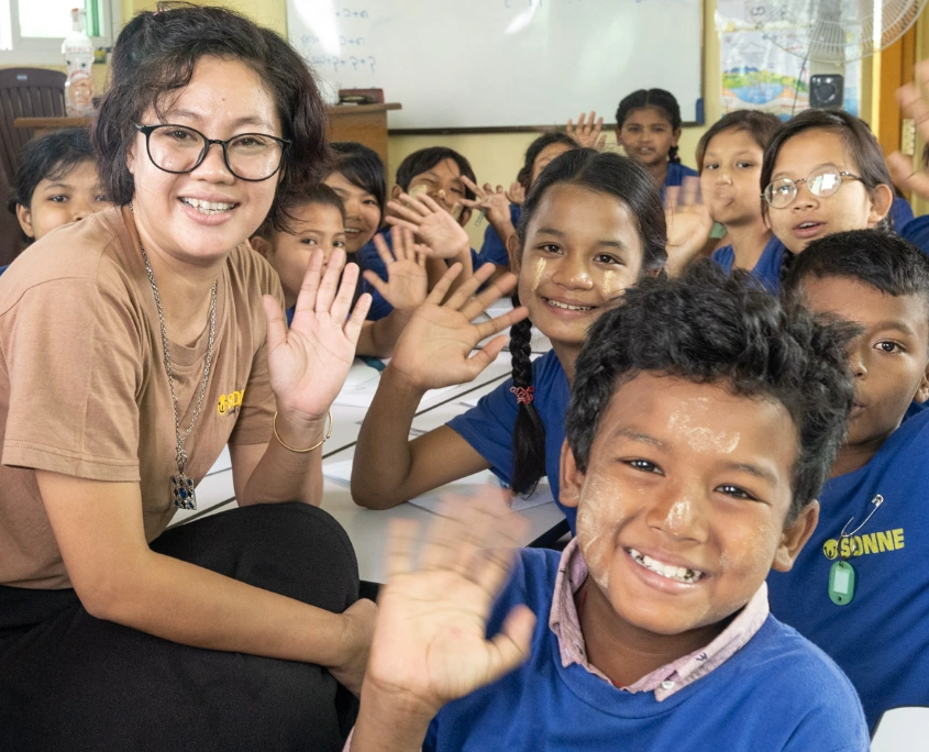 Lehrerin und mehrere Kinder in SONNE-Shirts sitzen gemeinsam im Klassenzimmer und winken freundlich in die Kamera in Yangon.