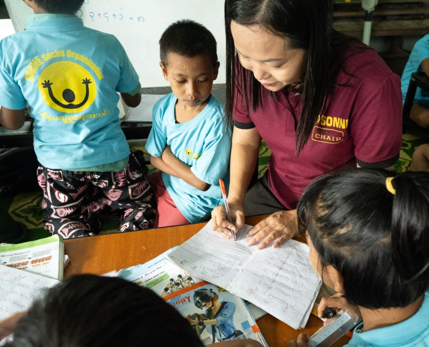 Mitarbeiterin von SONNE hilft mehreren Kindern beim Schreiben und Lernen an einem Tisch in einer Tagesbetreuungsstätte in Myanmar.