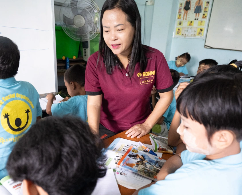 SONNE Lehrerin hilft mehreren Kindern beim Schreiben und Lernen an einem Tisch in einer Tagesbetreuungsstätte in Myanmar.