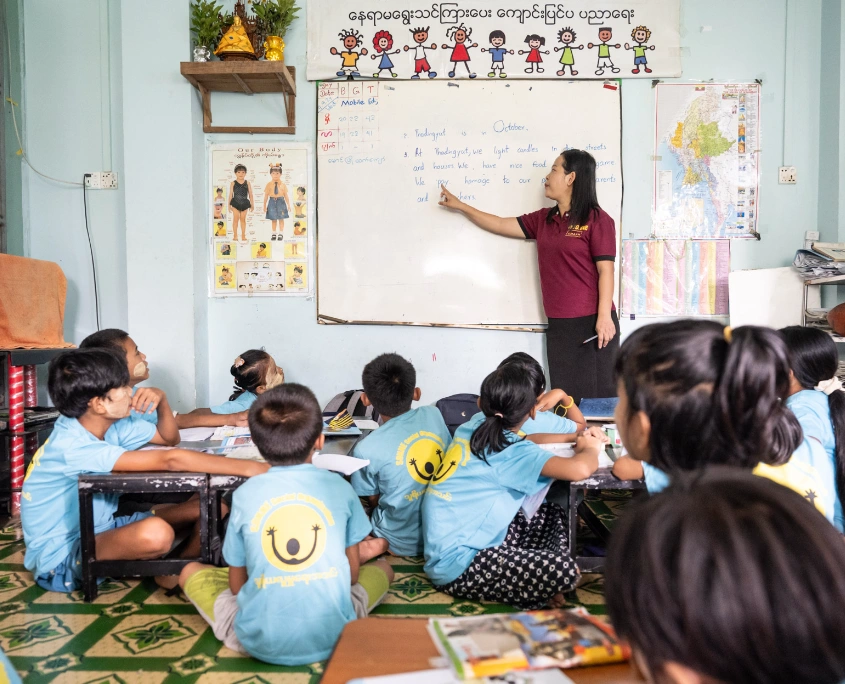 Lehrerin von SONNE steht vor einer Tafel und unterrichtet eine Gruppe von Kindern, die aufmerksam im Klassenzimmer in Yangon sitzen.